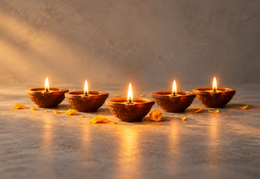 A neat row of small lit clay diyas on a stone surface, one diya slightly forward and glowing brighter, a few marigold petals scattered nearby